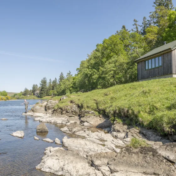A man fishing near the grassy bank of the River Dee, casting his line into the water, with a small wooden hut perched on the embankment behind him.